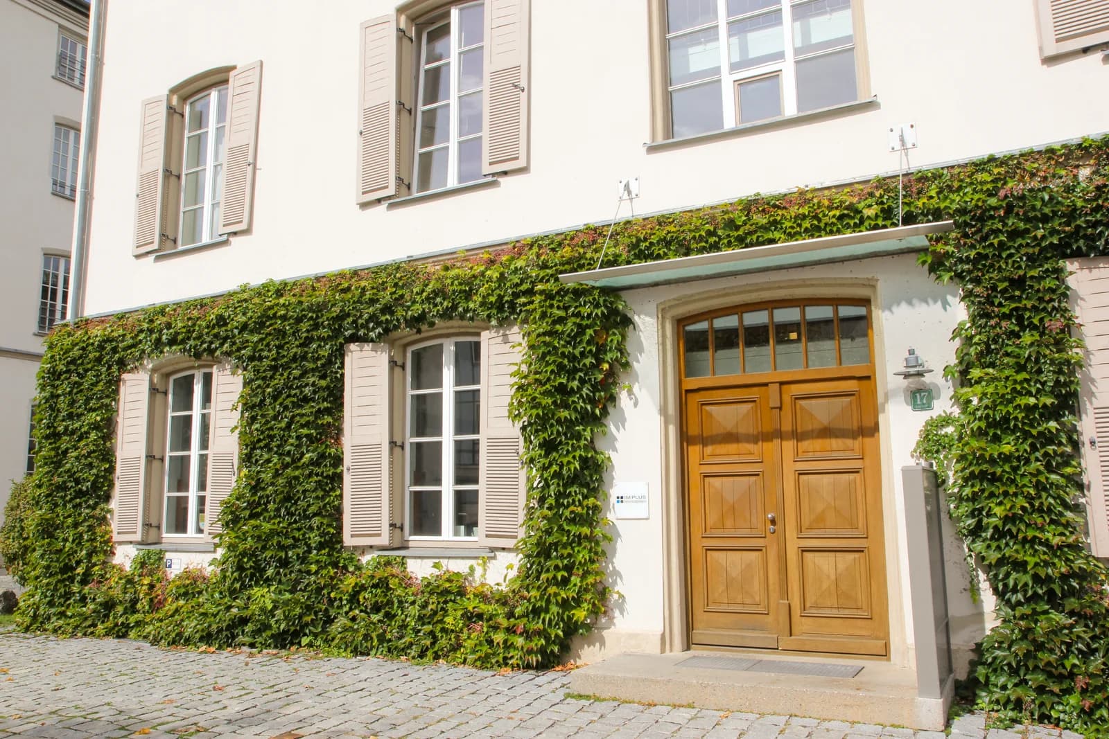 Der Eingang zum Büro: Holztür mit der Hausnummer 17 und dem Schild von IM PLUS Immobilien, daneben begrünte Fenster